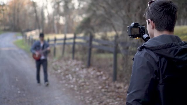 Using a gimbal, a teen filmmaker walks backwards filming a music video with a guitar player walkin.g with a wood fence around a barnyard 