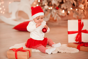 Obraz na płótnie Canvas Cute baby girl wearing santa hat holding Christmas ball in room over Christmas lights. Holiday season.