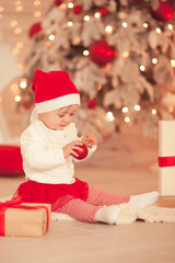 Obraz na płótnie Canvas Smiling baby girl 1 year old holding Christmas ball over lights background in room. Holiday season. Childhood.