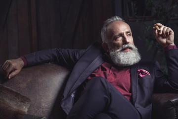 Portrait of smiling bearded old businessman keeping tobacco while locating on couch. Relax concept