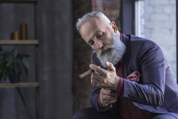 Portrait of serious unshaven pensioner buttoning cuff link on sleeve of elegant shirt while holding cigar in arm. Relax concept