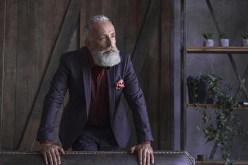 Portrait of thoughtful bearded pensioner leaning on sofa in living room. Work concept
