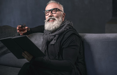 Low angle portrait of beaming bearded retire tasting delicious liquid while looking through volume...