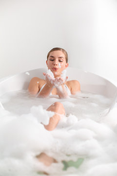 Full length portrait of cheerful young woman lying in water in bathtub and blowing foam with joy. Luxury concept