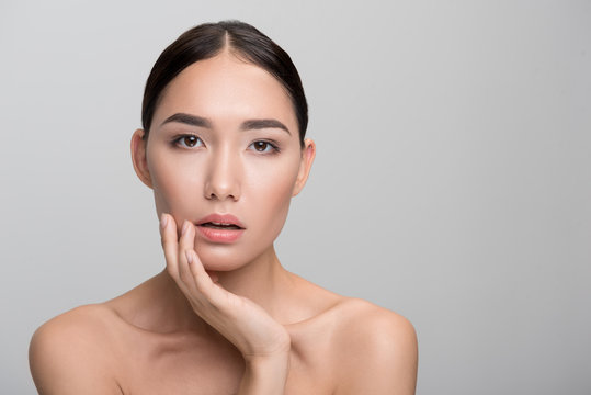 Beauty concept. Portrait of naked young asian woman is touching her cheek with her fingers while looking at camera seriously. She is taking pleasure from her fresh soft skin. Isolated with copy space