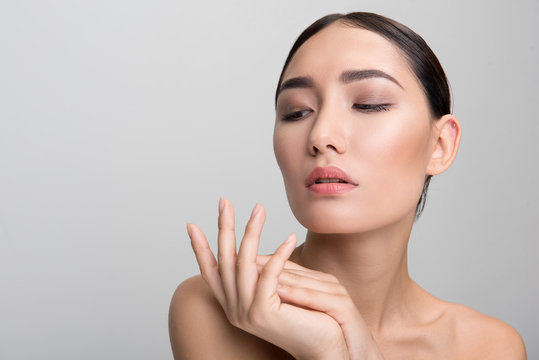 Skincare. Portrait of elegant naked dark-haired asian woman touching her palm with tenderness while being delighted with her skin and looking down dreamily. Isolated with copy space