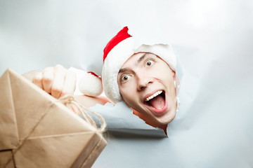 Christmas New Year concept, Stylish guy santa hat breaks through paper and looking for something with happy face and hold gift in hand, with place for text