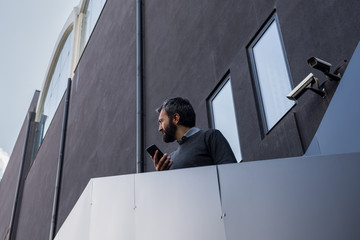 Fototapeta premium Young adult business man holding smartphone look left standing on black metal outside staircase on industrial black facade.