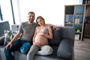 Full length portrait of joyful young family relaxing at home together. Man is embracing expectant woman and smiling while sitting on couch