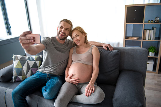 Portrait of happy married couple making selfie on smartphone and laughing. Pregnant woman is sitting on sofa and touching her bare abdomen with gentleness