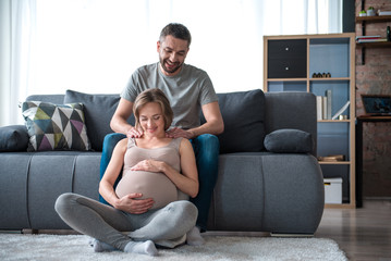 Just relax and get some pleasure. Full length portrait of glad future father doing massage for his expectant wife. Lady is sitting on floor and smiling