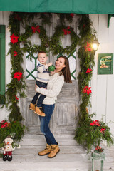Obraz na płótnie Canvas Happy mother with little son. Child boy in sweater staying on porch snow steps at light house with decorated in red green New Year door at home. Christmas good mood. Family and holiday 2018 concept