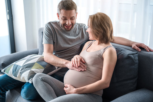 Baby is pushing. Portrait of excited married couple touching female pregnant abdomen and laughing. They are sitting on couch at home