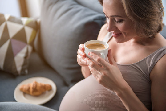 Enjoying Free Time Yet. Side View Of Cheerful Pregnant Woman Drinking Coffee While Relaxing On Sofa. She Is Looking Into Cup And Smiling. Copy Space