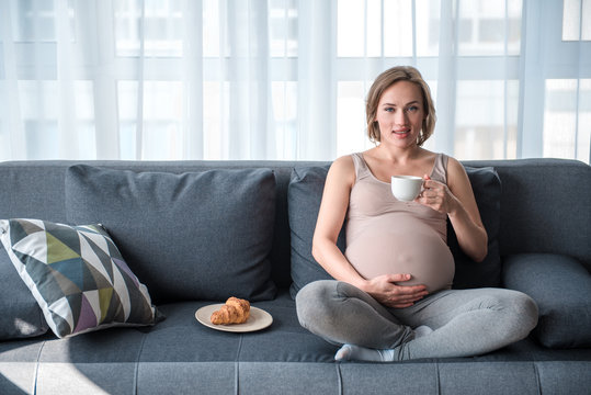 Full length portrait of glad expectant mother having breakfast in living room. She is touching her belly with gentleness and smiling. Copy space