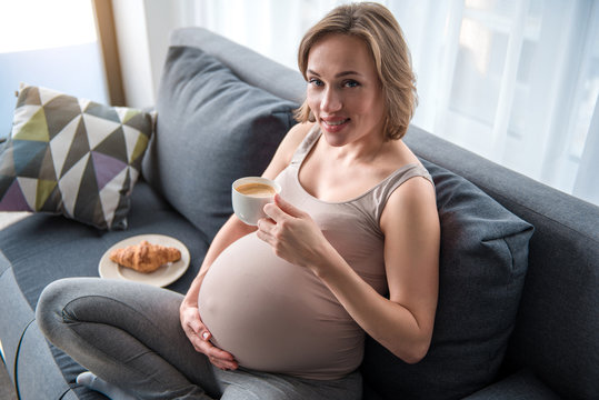 Good morning. Portrait of happy pregnant woman enjoying coffee with croissant at home. She is looking at camera and smiling