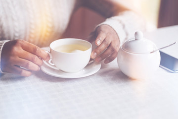 Hot tea. African woman wearing knitted sweater holding her hands on the table while going to drink tea