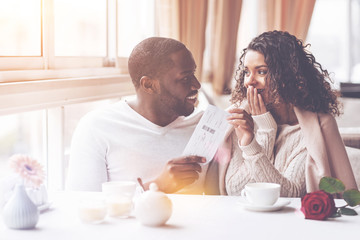 Love in the air. Smiling black male person turning his head to girlfriend while looking at her and giving ticket for trip