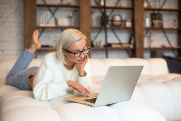Fototapeta premium Pretty young blonde girl working on a laptop sitting on the bed. Beautiful young woman in a fluffy white sweater and glasses working from home on a laptop lying on the bed in the room with white walls