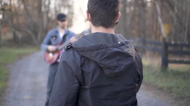 Using a gimbal, a teen filmmaker walks backwards filming a music video with a guitar player walking with a wood fence around a barnyard 