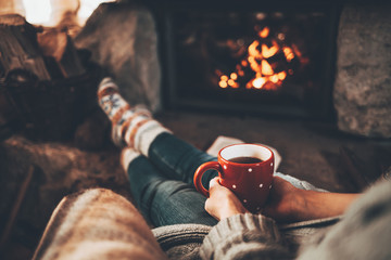 Woman in woollen socks by the fireplace. Unrecognisable relaxes by warm fire with a cup of hot...