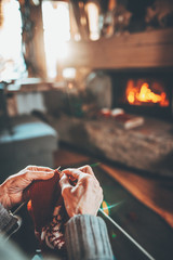 Senior woman hands knitting by the fireplace. Unrecognisable grandmother relaxes by warm fire making handmade gifts for her family. Cozy atmosphere. Winter and Christmas holidays concept.