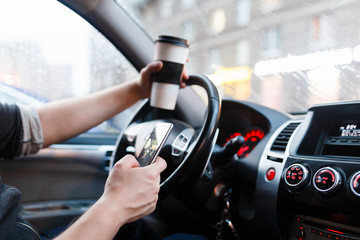 Stylish driver with a smartphone in hand and paper cup of hot coffee in the driver's seat. The concept of inattention at the wheel, rest, coffee break to cheer.