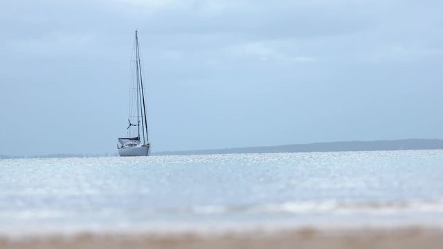 Shot of a yacht anchored on a blue cloudy day, with the beach present in the foreground and another shoreline in the background.