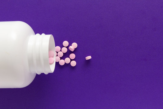 Group pink tablets. Capsules spilling out of white bottle. Purple background.