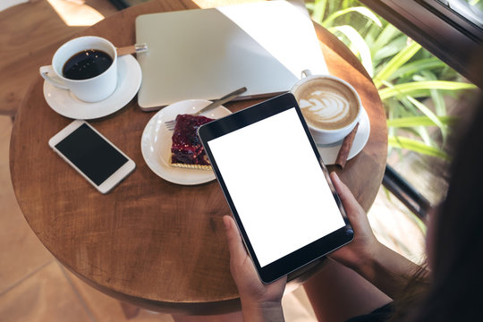 Top view mockup image of hands holding black tablet pc with blank white screen , smart phone , laptop , coffee cup and cake on wooden table in cafe