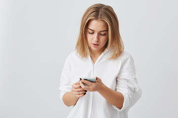 Fototapeta premium Serious young girl with fair hair wearing white shirt surfing the Internet on cell phone, looking at the screen with concentrated expression, checking email against gray copy space studio wall