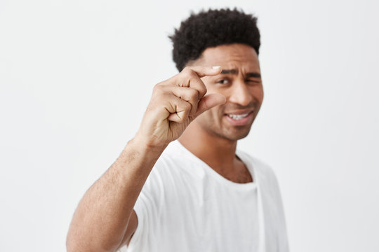 Copy space. Selective focus. Close up of young beautiful dark-skinned man with afro hair in white t-shirt gesticulating with hand, looking in camera with sarcastic face expression.