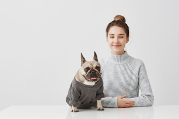 Indoor shot of joyful female pet lover with brown hair in bun. Housewife posing with her pedigree dog dressed in jumper sitting at table taking pleasure together. Positive human expressions