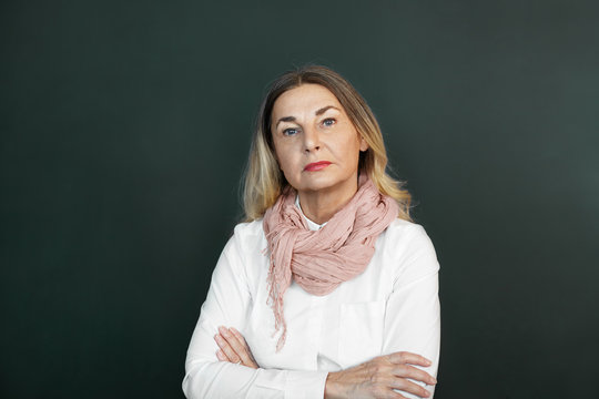Blonde mature woman with skeptical strict expression on her wrinkled face, posing in studio, keeping arms folded, expressing suspicion or disapproval. Negative human emotions, feelings, reaction
