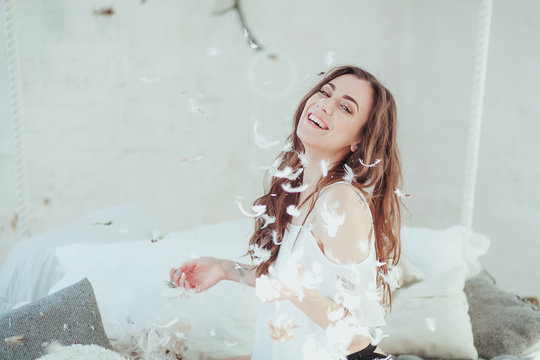 Beautiful young woman in a bed dropping the feathers and smiling. Close-up horizontal portrait.