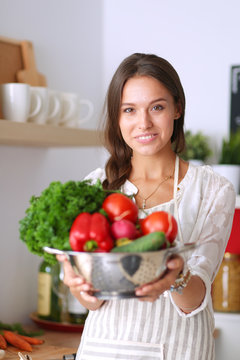 Smiling young woman holding vegetables standing in kitchen. Smiling young woman