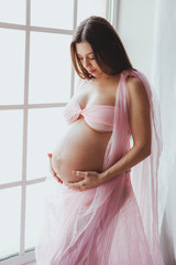Close-up portrait of tender pregnant beauty young woman standing in front of the window and touching her belly at home. Loft minimalistic interior