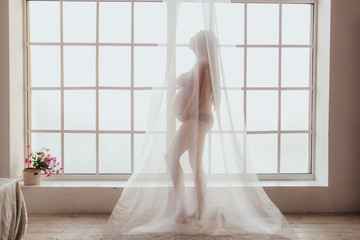 Beautiful young mom standing in front of window covered with chiffon curtain.