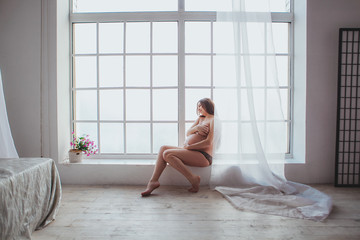 Pregnant woman in black underwear sitting on window sill. Young woman expecting a baby. Big window in loft interior