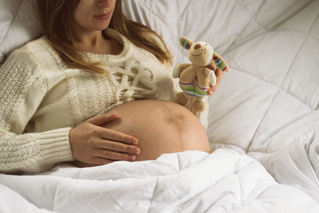 Pregnant young girl lying in bed holding a toy, the other hand on the abdomen