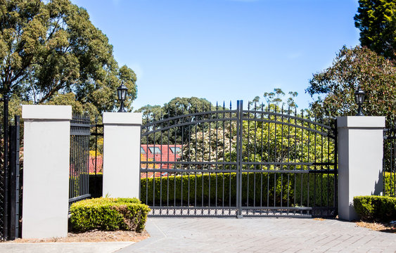 Black metal driveway entrance gates set in brick fence with garden trees in background