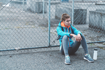 Athlete pretty woman with backpack, headphones preparing for running on the city street. Resting after jogging. Sitting with bottle of water. Sport tight clothes. Horizontal.