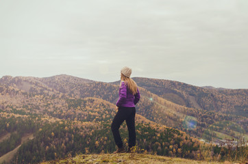 Naklejka na ściany i meble A girl in a lilac jacket looks out into the distance on a mountain, a view of the mountains and an autumnal forest by an overcast day. Free space for text