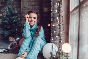 Beautiful woman sits at home near window with christmas lights. New Year.