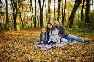Fototapeta premium Happy caucasian family of mom dad and little girl with boy sitting on plaid at majestic autumn fall forest.