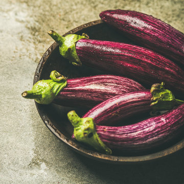 Fresh raw Fall harvest purple eggplants or aubergines in wooden bowl over grey concrete stone background, selective focus, square crop. Healthy Autumn vegan cooking ingredient
