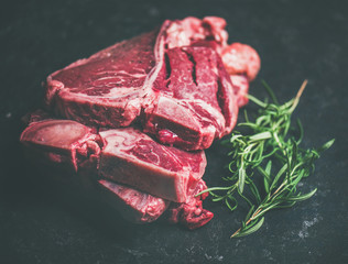 Close-up of fresh raw uncooked beef meat t-bone steaks placed in stack with rosemary over black stone background, selective focus