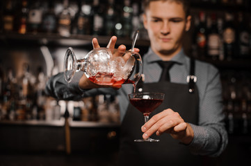 Young barman pouring alcoholic drink into the cocktail glass