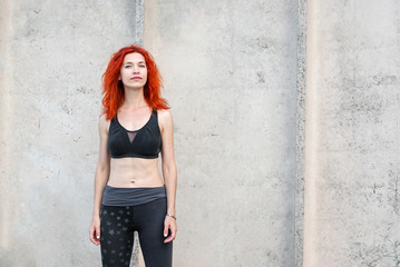 Pretty girl with red hair in sportswear stands on a gray background concrete wall