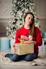 Portrait of beautiful woman with gift sitting by Christmas tree at home